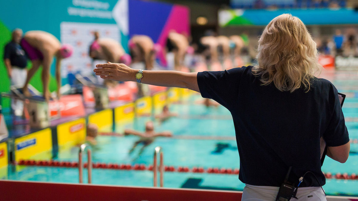 An official holds her arm out while swimmers are on the starting blocks.