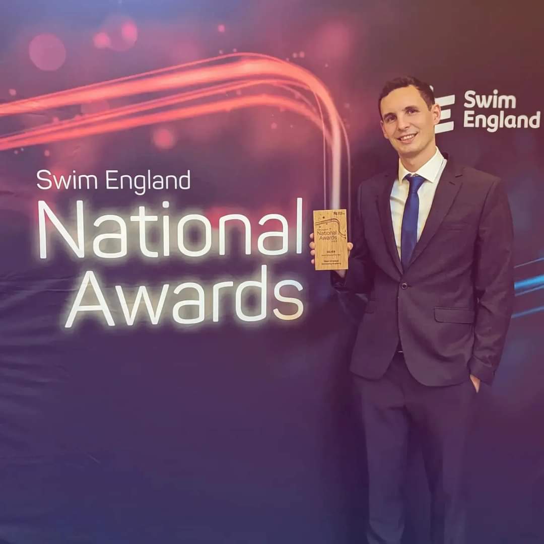 A person from WGSA Swim School standing in front of the National Awards banner holding a trophy.