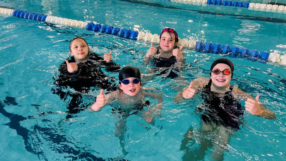 4 swimmers are in an indoor swimming pool, positioned near a blue-and-white lane divider. Each swimmer is wearing a swim cap and goggles, with heads and shoulders above the water, and all are giving a thumbs-up gesture toward the camera. The clear blue pool water and tiled pool edge are visible in the background.